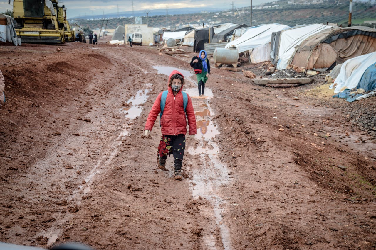 Children in a muddy refugee camp in Idlib, Syria during a winter day, facing harsh conditions.